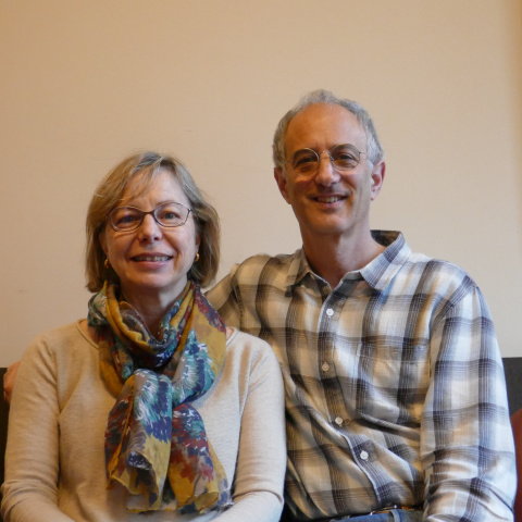 Photo of Bill and Holly Marklyn seated side by side, smiling at the camera; Holly wears glasses and a colorful scarf, and Bill wears a plaid shirt, against a simple indoor background.