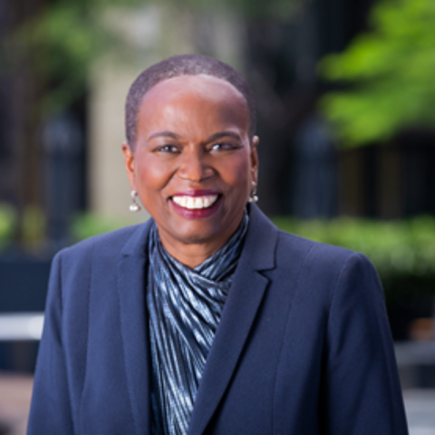 Headshot of Judy Belk smiling, wearing a navy blazer and patterned blouse, with a blurred outdoor background of greenery and buildings.