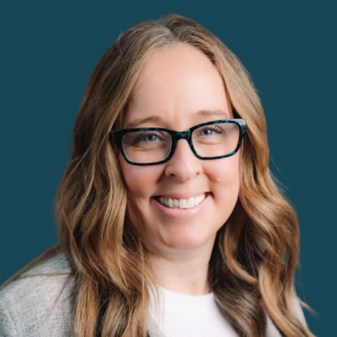 Headshot of Karri Matau smiling, wearing glasses with long wavy hair against a solid teal background