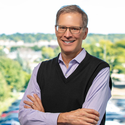 Headshot of Kevin Walker smiling with arms crossed, wearing glasses, a light purple shirt and a black sweater vest, with a bright outdoor background of trees and a parking area.