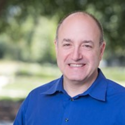 Headshot of Larry Kramer smiling, wearing a bright blue collared shirt, with a blurred outdoor background of trees and greenery.