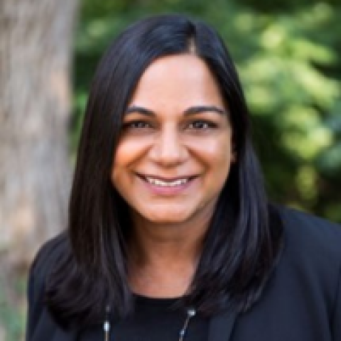 Headshot of Monika Kalra Varma smiling, wearing a black jacket with a blurred outdoor background of trees.