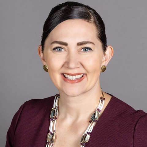 Headshot of Nichole June Maher smiling, wearing a burgundy top, gold earrings and a beaded necklace, against a plain gray background.