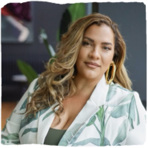 Headshot of Esperanza Tervalon smiling softly, wearing large gold hoop earrings and a patterned blazer with green leaf designs, seated indoors with plants and artwork in the background.