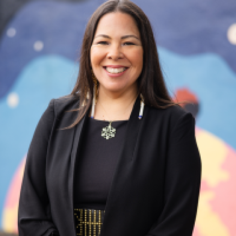 Headshot of Se-ah-dom Edmo smiling, wearing a black outfit with a beaded belt and earrings, standing in front of a colorful outdoor mural.