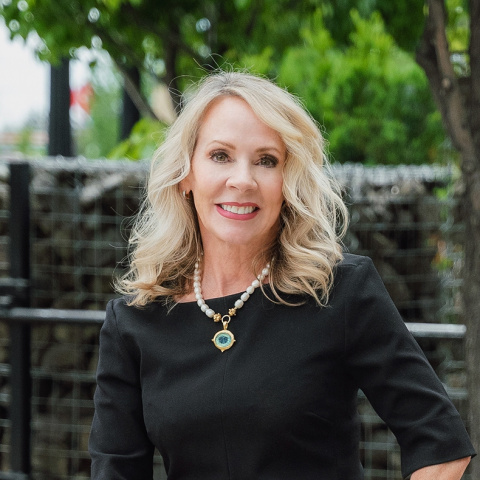 Headshot of Sharon Miracle smiling, wearing a black dress and a pearl necklace with a pendant, standing outdoors with trees and a metal fence in the background.