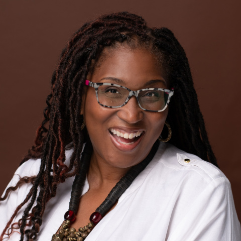 Headshot of Tanya Edelin smiling, wearing patterned glasses, a white top and layered jewelry, against a solid brown background.