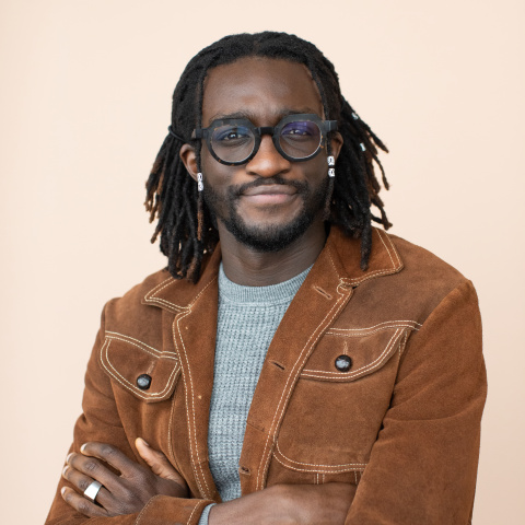 Headshot of Juma Sei smiling with arms crossed, wearing glasses, a brown suede jacket and a gray shirt against a soft peach background.