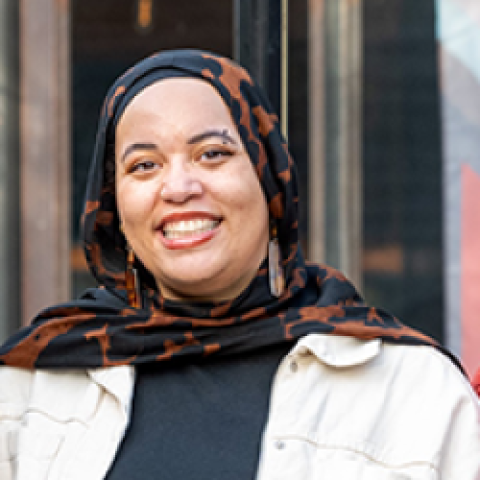 Aisha Al-Amin Headshot, wearing a brown and black hijab, black shirt and white jacket, smiling in front of a blurred background