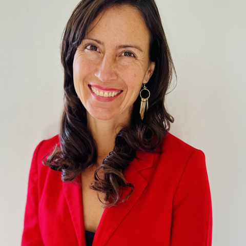 Pearl Lujan headshot, with brown shoulder length hair and a red shirt, smiling in front of a white background