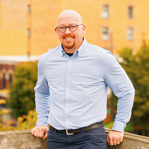 Headshot of Seth Kirby wearing a blue dress shirt in front of a yellow building outdoors, smiling