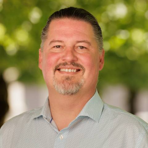 Zeke Smith headshot, Zeke is smiling with short brown hair, wearing a light grey shirt in front of a blurred background
