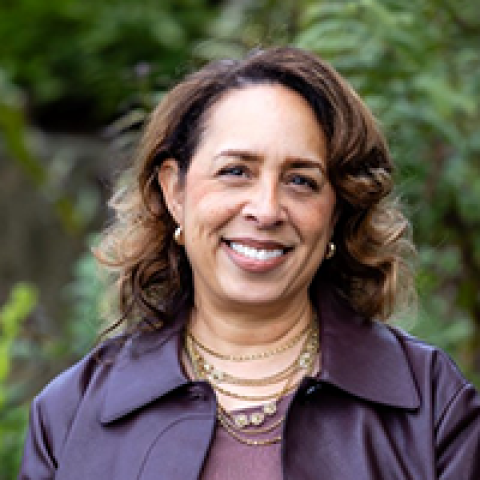 Headshot of a smiling Maya Thornell-Sandifor with medium skin tone and shoulder-length wavy brown hair, wearing layered gold necklaces and a purple jacket, photographed outdoors against a softly blurred green background.
