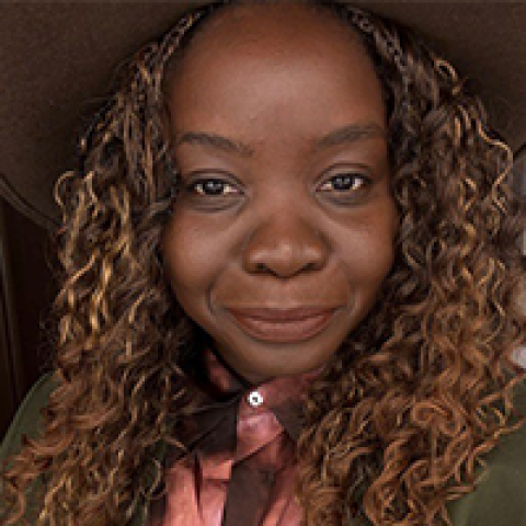 Headshot of a smiling Valériana Chikoti-Bandua with medium-dark skin and long, tightly curled hair with warm highlights. She wears a satin pink blouse under a dark jacket, photographed indoors against a softly shadowed background.