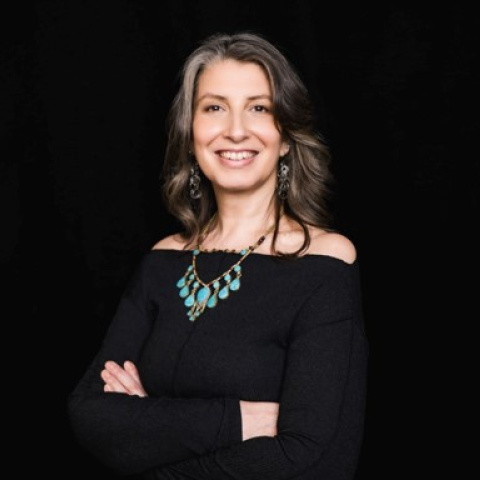 Headshot of M. Gabriela Alcalde, smiling with shoulder-length dark hair and wearing a black off-the-shoulder top, turquoise statement necklace and dangling earrings, with her arms crossed against a black background.