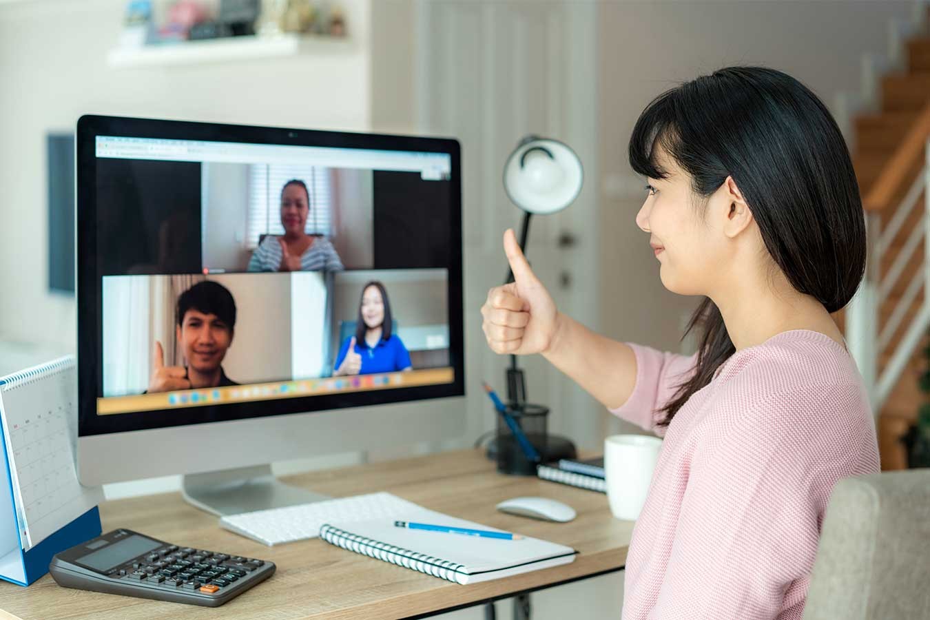 Image of an Asian woman working from home giving a thumbs up sign to videoconference colleagues visible on her laptop 