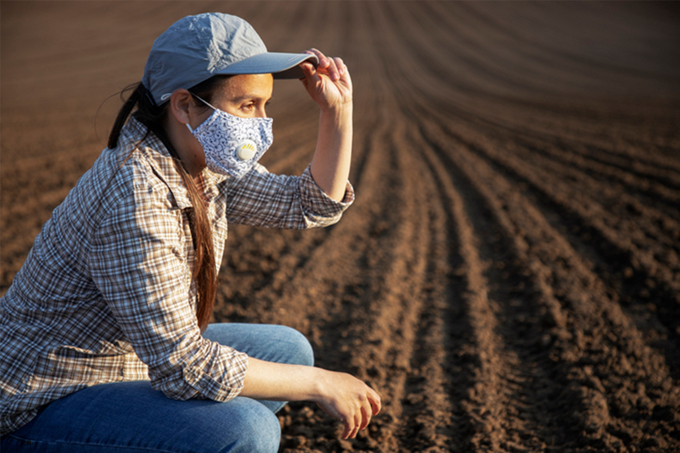 Woman wearing  blue cap, a flannel, jeans and a mask squatting in a rural dirt field and looking off in the distance.