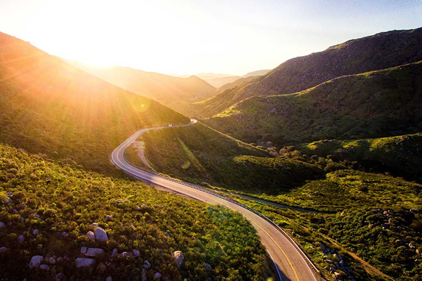 Picture of sunshine breaking over hills into a rural valley with a two lane winding road