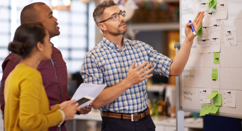 A team of professionals collaborating in an office environment. A man in a checkered shirt explains ideas while pointing to a whiteboard covered with sticky notes and pinned cards.