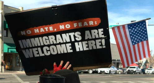 Photo of a protest sign reading, “No hate, no fear: Immigrants are welcome here!” held up on a city street, with an American flag and parked cars in the background.