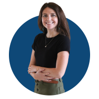 Headshot of Audrey against a blue background. Audrey wears a black short-sleeve top, olive-green high-waist pants, a pendant necklace, and teal drop earrings.