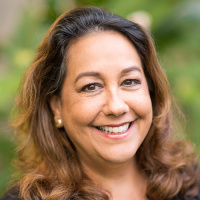 Headshot of Michelle Kauhane smiling with wavy brown hair and warm brown eyes, wearing small gold stud earrings, photographed outdoors against a softly blurred green background.