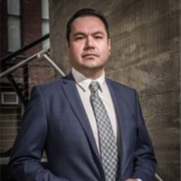 Headshot of Brian Tanner, looking at the camera with a neutral expression, wearing a navy suit and patterned tie against an urban concrete and brick background.