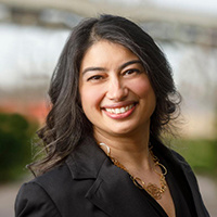 Headshot of Kaberi Banerjee Murthy, smiling, with shoulder-length dark hair, wearing a black blazer and gold necklace against a softly blurred outdoor background.