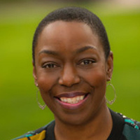 Headshot of Shona Carter, smiling, with short hair, wearing hoop earrings and a patterned top against a blurred green outdoor background.