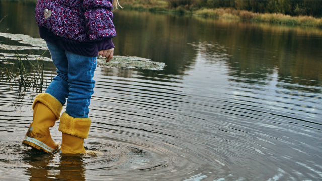 A child in yellow rubber boots stands in the water, blue jeans and a purple jacket, with her feet under water in a flooded street.