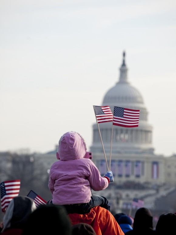Image of a child on parent's shoulders waving a small US flag in a crowd of people in front of the U.S. capitol