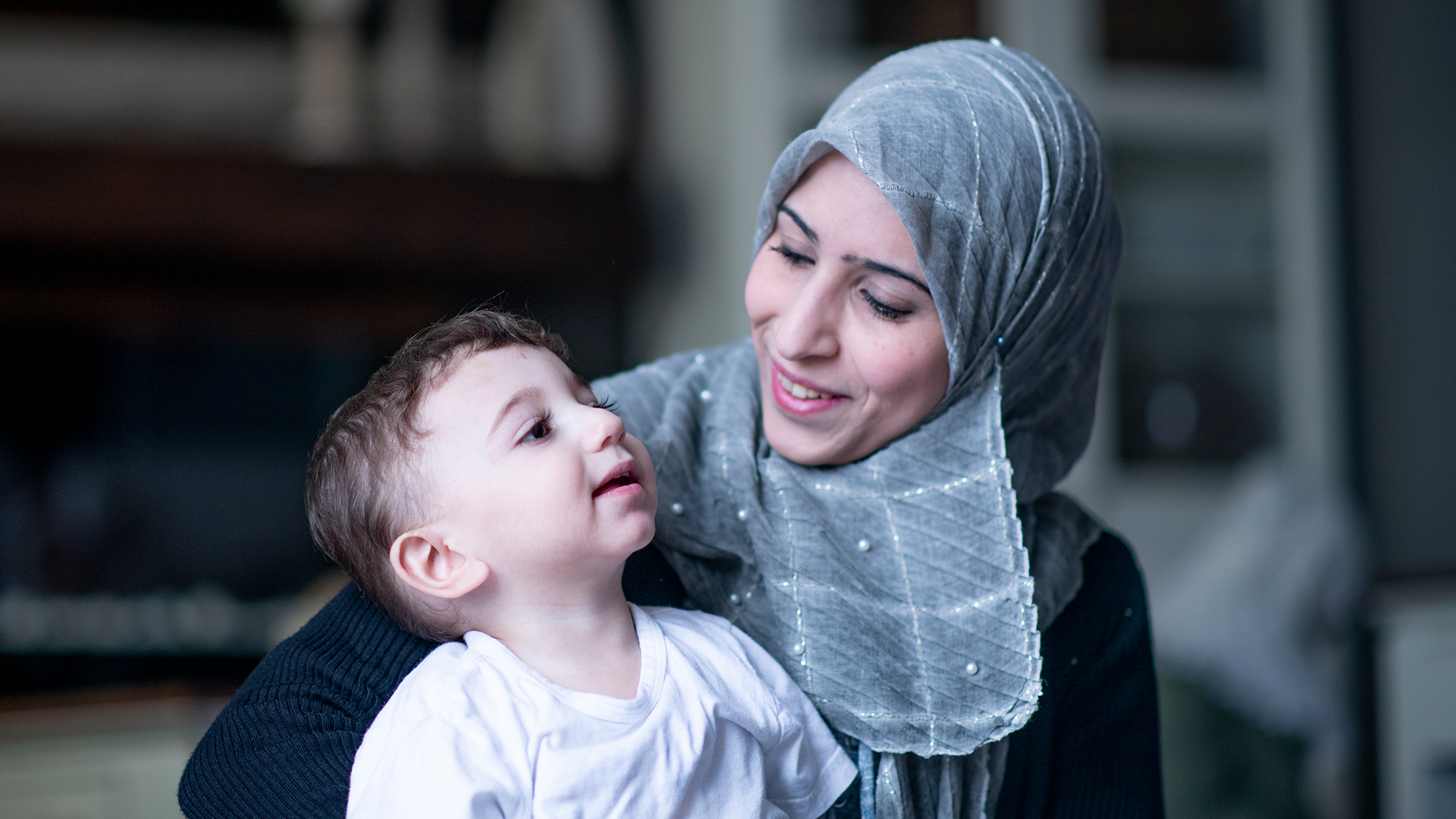 Picture of Muslim mother wearing blue-grey hijab looking lovingly at toddler son in her lap wearing white shirt