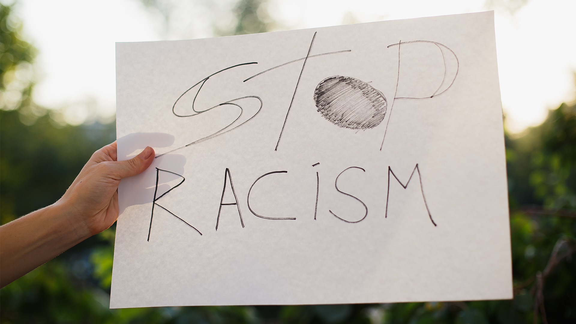 Woman holding white paper with text "Stop racism". Against the background of trees and sunlight