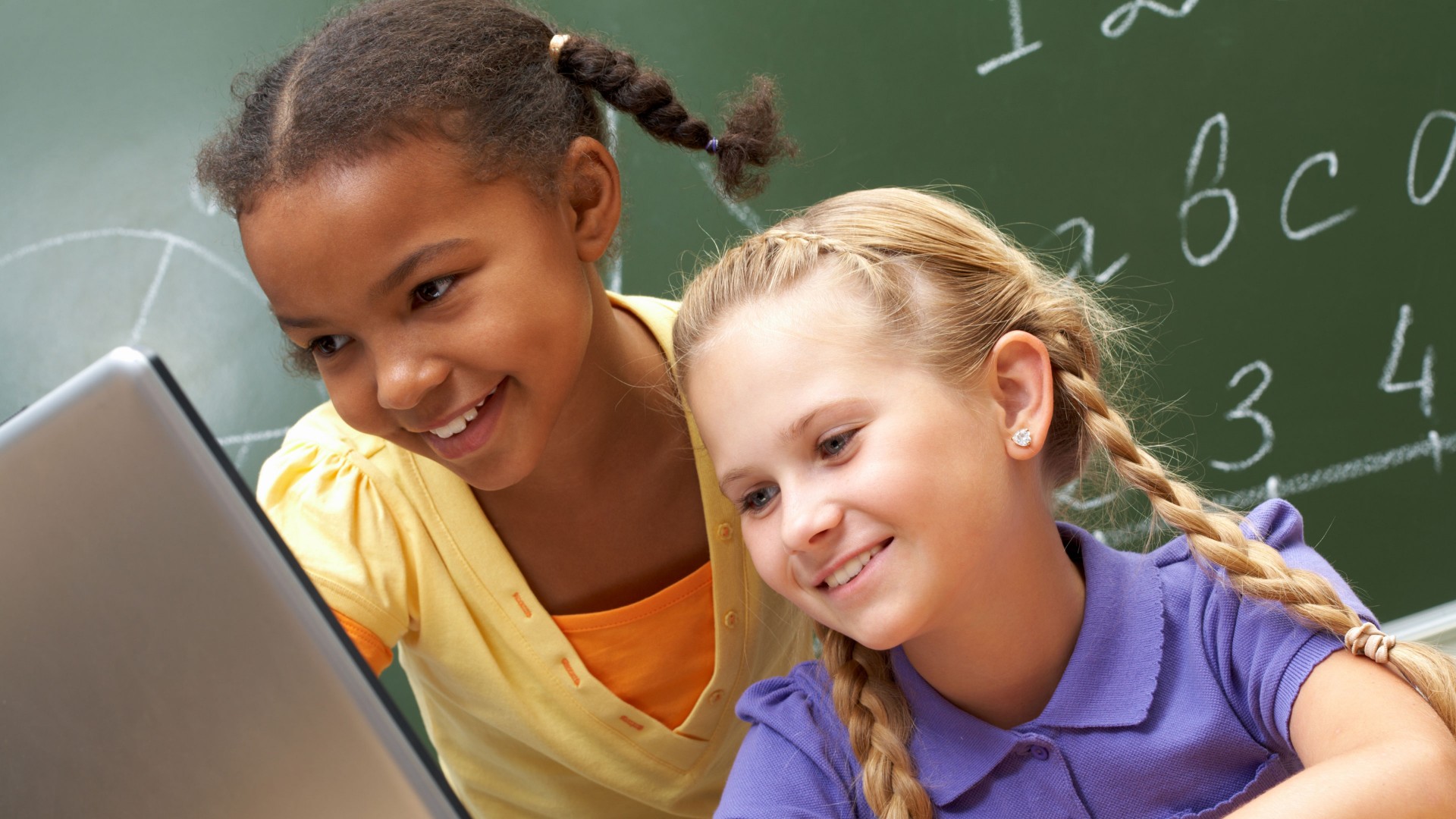 Two smiling school-age girls, one black, one white, loot at a laptop with a blackboard in the background