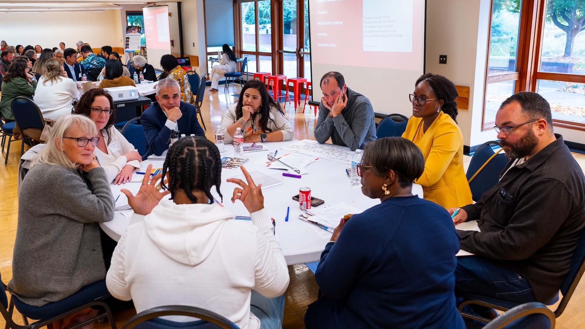 A diverse group of people sits around a round table engaged in a focused conversation during a workshop. The group includes men and women of different ages and backgrounds, with materials and papers spread across the table. A presentation screen in the ba