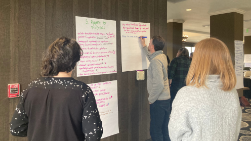 Several people are standing in front of large sheets of paper taped to a wall, reviewing and writing notes during a collaborative session. The papers contain handwritten headings and bullet points, including “3. Request for Proposals.” The setting is an indoor meeting space with neutral walls and patterned carpet at Philanthropy Northwest’s Grantmaking with an Equity Lens (GEL) workshop in Eugene, Oregon.