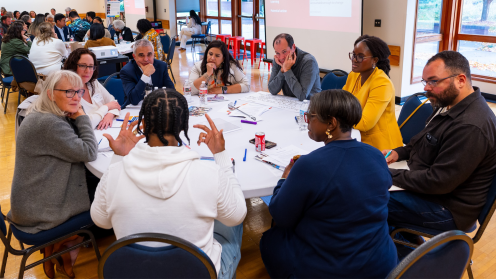 Photo of a roundtable discussion at the “Trust-Based Philanthropy in South King County” gathering, with participants seated in a bright meeting room collaborating on worksheets and notes. A presentation slide is projected in the background, and other groups are visible working at tables throughout the space.