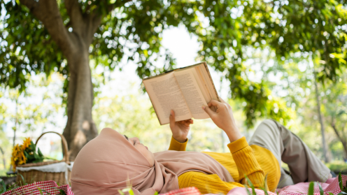 Photo of a young Muslim woman reading a book on a red checkered blanket with a picnic basket with yellow sunflowers nearby. She is surrounded by green trees.