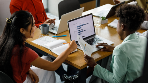 High angle view of two young businesswomen reviewing a document and data on laptop during a meeting with their colleagues.