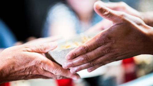 Hands of two people with different skin tones meet as one passes a bowl of food to the other. The close-up focuses on the exchange, conveying care, generosity, and shared responsibility, with the background softly blurred to emphasize the act of giving.