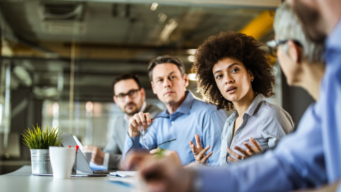 A woman with voluminous curly hair and medium-dark skin speaks and gestures during a meeting while three colleagues listen attentively. A laptop, coffee cup, and small potted plant sit on the table, the glass-walled office suggesting a focused group discussion.