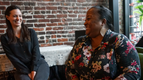 Two women sit facing each other in conversation, smiling and laughing together. On the left, a light-skinned woman with long dark hair wears a dark button-down shirt. On the right, a woman with medium-dark skin and hair pulled back wears a black floral top and large earrings. They sit in a warm, informal setting with an exposed brick wall behind them, conveying connection, ease, and shared dialogue.