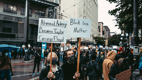 Two protesters wearing face masks stand in a city street holding signs that read “Ahmaud Arbery, Breonna Taylor, George Floyd” and “Black Lives Matter.” A larger crowd fills the background between downtown buildings on a rainy day, many carrying signs, during a racial justice demonstration.
