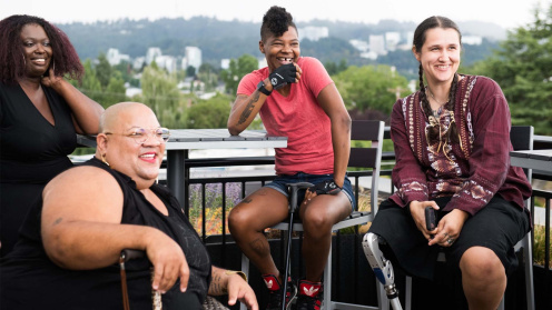 Four disabled people of color with canes and prosthetic legs laugh while chatting. They are on a rooftop deck, in chairs of various height, with greenery and city high-rises in the background.