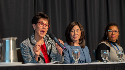 A photo of Elizabeth Ripley speaking into a handheld microphone at a panel discussion. She wears red glasses, a patterned black-and-white blazer and a red blouse, gesturing as she speaks. Two panelists sit to her right, listening; a table with water glasses and a dark stage curtain form the backdrop.