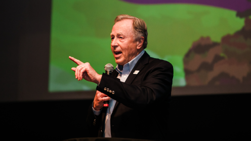 Mike Halligan speaks at the PNW24 Conference in Missoula, Montana, holding a microphone and gesturing toward the audience. He wears a dark blazer over a light blue shirt, standing in front of a large green and purple projection screen.