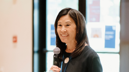 Photo of Jill Nishi smiling while speaking into a handheld microphone at an indoor event, with bright windows in the background.