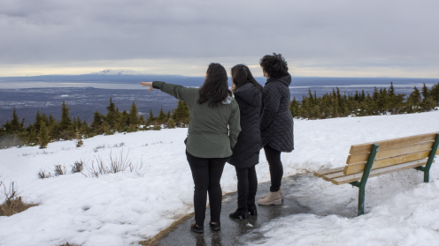 Three people stand on a snowy overlook in Alaska, looking out toward mountains and water under a cloudy sky. One person points toward the view, with evergreen trees and a wooden bench nearby.