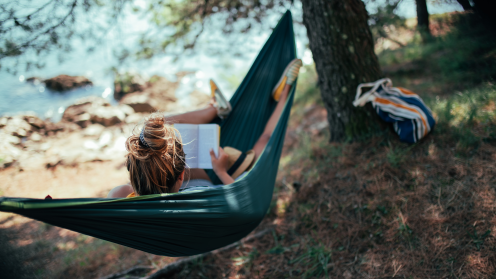 A person relaxes in a dark green hammock, reading a book beneath trees near a rocky shoreline. A striped bag sits nearby, and sunlight filters through the trees onto the water and forest floor.