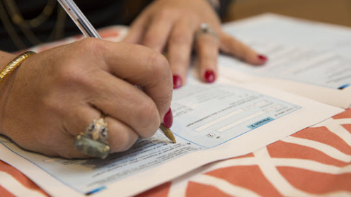 Close-up photo of a person filling out a paper form with a pen, with documents spread across a patterned tablecloth.